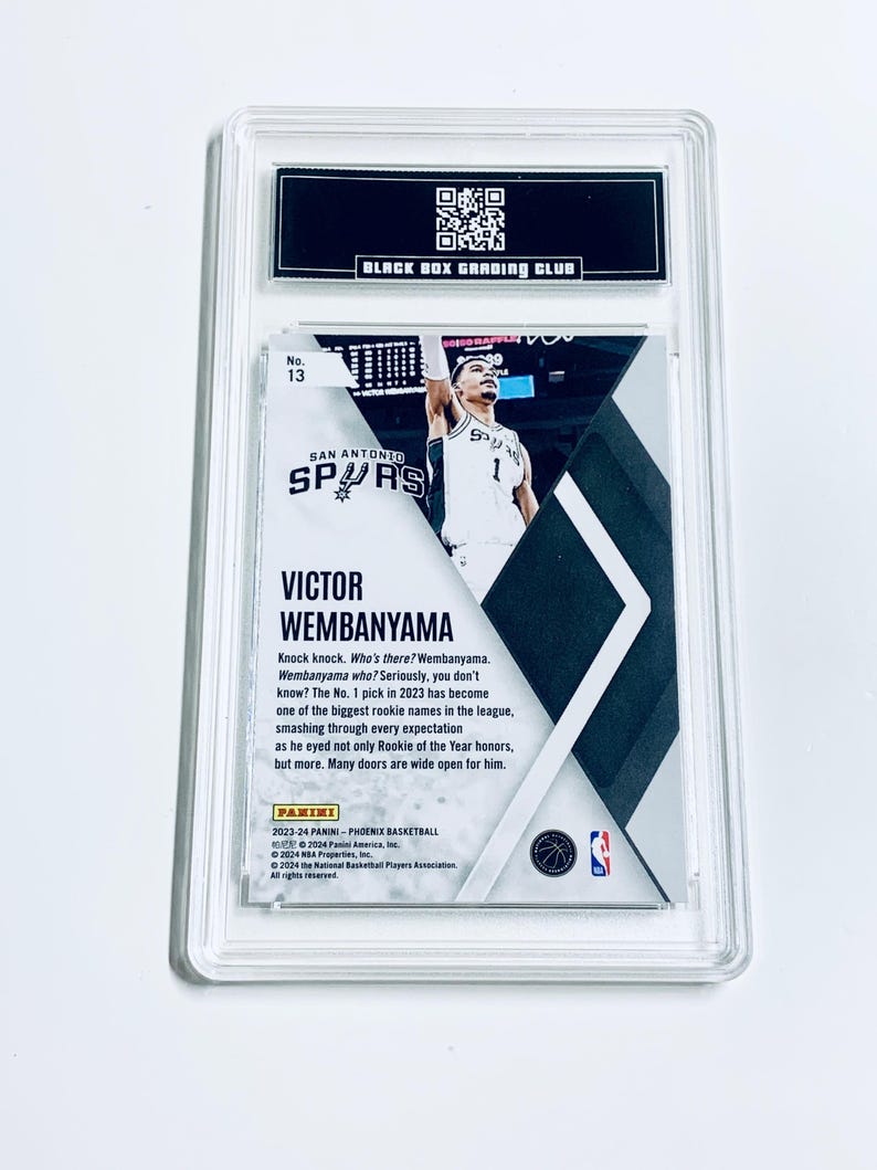 K&ouml;nnte beinhalten: Eine bewertete Basketballkarte mit Victor Wembanyama von den San Antonio Spurs. Die Karte ist in einem transparenten Kunststoffhalter mit dem Text "BLACK BOX GRADING CLUB" oben eingeschlossen. Die Karte hat unten den Text "PANINI".