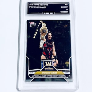 May include: A Topps Now WWE trading card featuring Stephanie Vaquer holding a championship belt. The card is encased in a clear protective holder. Text on the card includes "2025 TOPPS NOW WWE", "STEPHANIE VAQUER", and "LA PRIMERA, LA MEJOR: CAPTURES WOMEN'S WORLD TITLE".