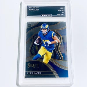 May include: A Puka Nacua football trading card from 2023, presented in a protective case. The card shows a player in a blue and yellow uniform, with the text "SELECT RAMS" and "PUKA NACUA". The case includes "BLACK BOX" and grading details.