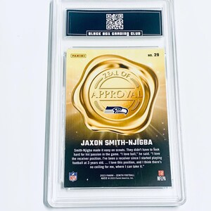 K&ouml;nnte beinhalten: Eine Panini Zenith Football-Sammelkarte von Jaxon Smith-Njigba, bewertet von Black Box Grading Club. Die Karte zeigt ein Goldfoliensiegel mit den Worten "Zeal of Approval" und dem Logo der Seattle Seahawks. Die Karte ist in einer transparenten Schutzh&uuml;lle eingeschlossen.