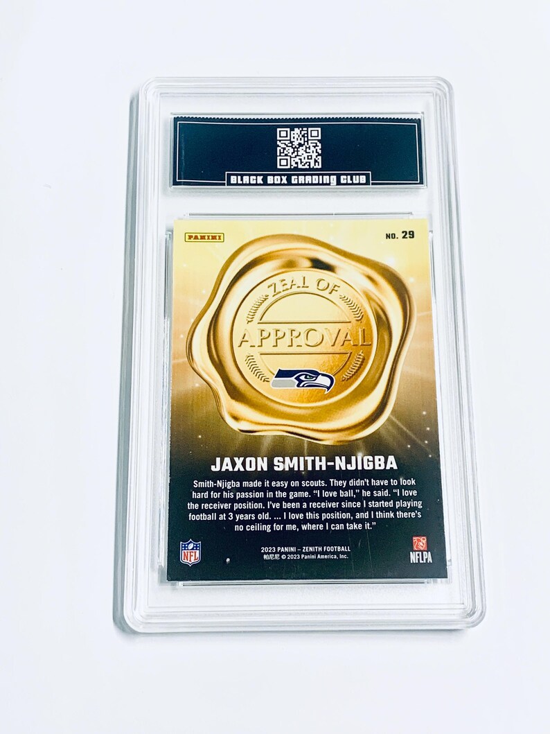K&ouml;nnte beinhalten: Eine Panini Zenith Football-Sammelkarte von Jaxon Smith-Njigba, bewertet von Black Box Grading Club. Die Karte zeigt ein Goldfoliensiegel mit den Worten "Zeal of Approval" und dem Logo der Seattle Seahawks. Die Karte ist in einer transparenten Schutzh&uuml;lle eingeschlossen.