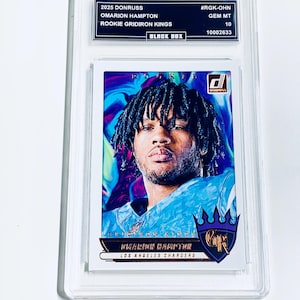 May include: A Donruss trading card of Omarion Hampton, a rookie for the Gridiron Kings, in a protective case. The card features a portrait of Hampton in a blue jersey against a colorful background. The card is labeled "Los Angeles Chargers."