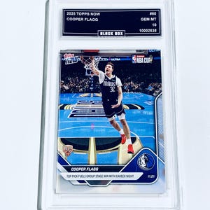 May include: A Topps Now basketball trading card featuring Cooper Flagg in action, graded GEM MT 10. The card is encased in a protective holder, with text including "2025 Topps Now" and "Black Box". The card shows Flagg in a blue and white uniform.