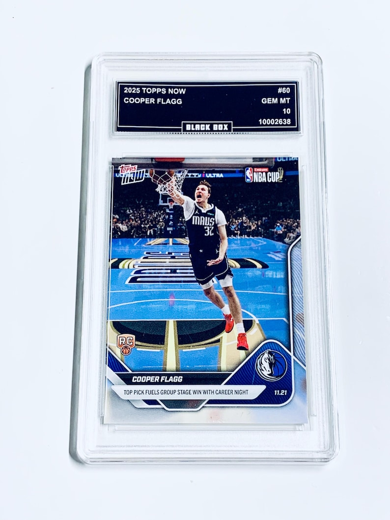 May include: A Topps Now basketball trading card featuring Cooper Flagg in action, graded GEM MT 10. The card is encased in a protective holder, with text including "2025 Topps Now" and "Black Box". The card shows Flagg in a blue and white uniform.