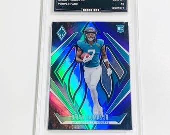 May include: A 2024 Phoenix trading card featuring Brian Thomas Jr. in a Jacksonville Jaguars uniform. The card has a purple fade design with the text "Purple Fade" and "Black Box". The card is graded GEM MT 10.