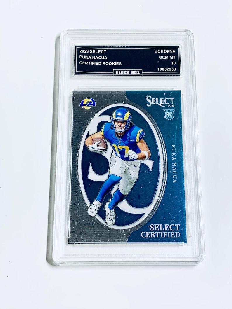 May include: A graded trading card featuring Puka Nacua in a blue and yellow uniform. The card is labeled "2023 Select Certified Rookies" and is encased in a protective holder. The card has the text "SELECT CERTIFIED" and "PUKA NACUA".