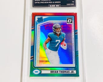 May include: A Brian Thomas Jr. football trading card from the 2024 Donruss Optic series. The card features a player in a Jacksonville Jaguars uniform, with red and green accents. The card is graded GEM MT 10 and encased in a protective holder.