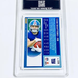 May include: A football trading card encased in a clear plastic holder. The card features a player in a blue uniform holding a football. The card's back includes player stats and the text "CAM SKATTEBO &bull; RB". The card is graded by "BLACK BOX GRADING CLUB".
