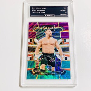 May include: A Bron Breakker wrestling card from the 2024 Select WWE series. The card features a shirtless wrestler in black shorts, set against a vibrant, multi-colored background. The card is encased in a protective holder, with text indicating it's a Tri-Color Prizm.