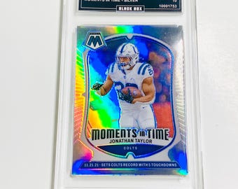 May include: A graded 2024 Mosaic Jonathan Taylor football card. The card features a player in a Colts uniform with the text "MOMENTS IN TIME". The card is encased in a clear protective holder. The card also has the text "11.21.21 - SETS COLTS RECORD WITH 5 TOUCHDOWNS".