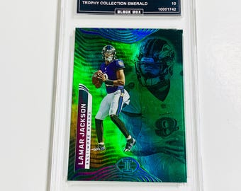 May include: A Lamar Jackson Baltimore Ravens trading card from the 2022 Illusions Trophy Collection Emerald set. The card is graded GEM MT 10 and encased in a protective holder. The card features a green and purple design.