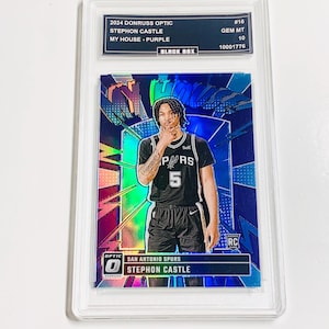 May include: A Stephon Castle basketball card from the 2024 Donruss Optic series. The card features a photo of the player in a black and white San Antonio Spurs uniform. The card is graded GEM MT 10 and encased in a protective holder.