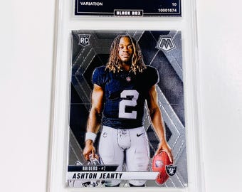 May include: A graded Ashton Jeanty football card from the 2025 Mosaic series. The card features a player in a Raiders uniform, holding a football. The card is encased in a clear protective holder.