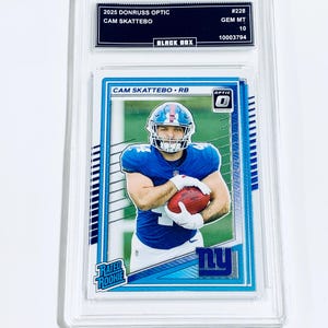 May include: A Cam Skattebo football card from the 2025 Donruss Optic series, graded GEM MT 10. The card features a player in a blue uniform holding a football. The card is encased in a protective holder.