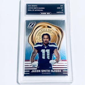 K&ouml;nnte beinhalten: Eine 2023 Zenith Jaxon Smith-Njigba Football-Sammelkarte, bewertet mit GEM MT 10, in einer Schutzh&uuml;lle. Die Karte zeigt einen Spieler im blau-wei&szlig;en Seahawks-Trikot mit der Nummer 11. Die Karte tr&auml;gt die Aufschrift "Zeal of Approval".