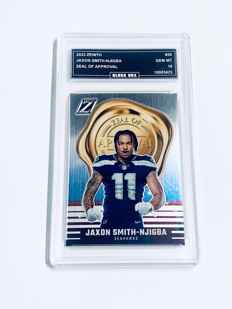 K&ouml;nnte beinhalten: Eine 2023 Zenith Jaxon Smith-Njigba Football-Sammelkarte, bewertet mit GEM MT 10, in einer Schutzh&uuml;lle. Die Karte zeigt einen Spieler im blau-wei&szlig;en Seahawks-Trikot mit der Nummer 11. Die Karte tr&auml;gt die Aufschrift "Zeal of Approval".