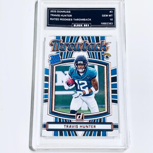 May include: A Travis Hunter football trading card from the 2025 Donruss set. The card features a player in a teal and black uniform with the number 12. The card is graded GEM MT 10 and encased in a protective holder.
