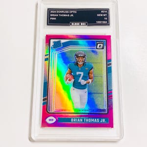 Puede incluir: Una tarjeta coleccionable rosa Donruss Optic 2024 con Brian Thomas Jr. de los Jacksonville Jaguars. La tarjeta est&aacute; calificada GEM MT 10 y est&aacute; dentro de un soporte protector. La tarjeta tiene el texto "RATED ROOKIE" y "OPTIC".