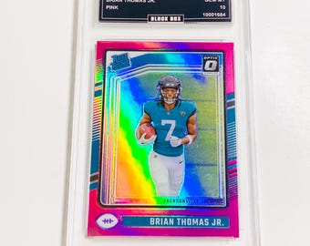 May include: A pink 2024 Donruss Optic trading card featuring Brian Thomas Jr. of the Jacksonville Jaguars. The card is graded GEM MT 10 and encased in a protective holder. The card has the text "RATED ROOKIE" and "OPTIC".