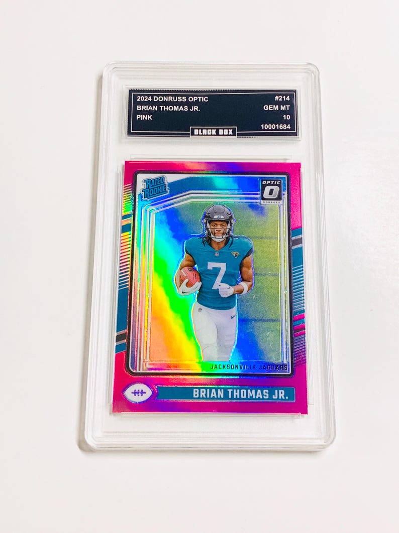 Puede incluir: Una tarjeta coleccionable rosa Donruss Optic 2024 con Brian Thomas Jr. de los Jacksonville Jaguars. La tarjeta est&aacute; calificada GEM MT 10 y est&aacute; dentro de un soporte protector. La tarjeta tiene el texto "RATED ROOKIE" y "OPTIC".