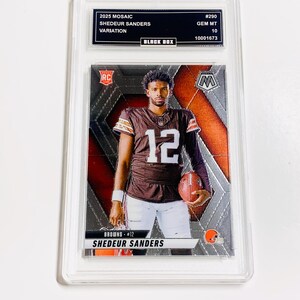 Puede incluir: Una tarjeta coleccionable graduada con Shedeur Sanders en un uniforme marr&oacute;n y blanco de los Browns, sosteniendo un bal&oacute;n de f&uacute;tbol. La tarjeta es del set 2025 Mosaic, con el texto "SHEDEUR SANDERS" y "BROWNS #12" visibles. La tarjeta est&aacute; dentro de un protector transparente.