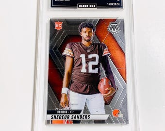 May include: A graded trading card featuring Shedeur Sanders in a brown and white Browns uniform, holding a football. The card is from the 2025 Mosaic set, with the text "SHEDEUR SANDERS" and "BROWNS #12" visible. The card is encased in a clear protective holder.