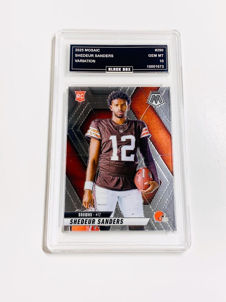 Puede incluir: Una tarjeta coleccionable graduada con Shedeur Sanders en un uniforme marr&oacute;n y blanco de los Browns, sosteniendo un bal&oacute;n de f&uacute;tbol. La tarjeta es del set 2025 Mosaic, con el texto "SHEDEUR SANDERS" y "BROWNS #12" visibles. La tarjeta est&aacute; dentro de un protector transparente.