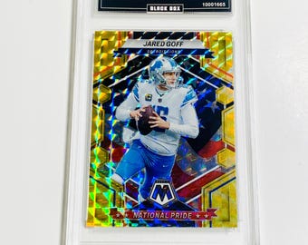 May include: A collectible trading card featuring Jared Goff of the Detroit Lions. The card has a mosaic design with yellow and blue accents. The card is encased in a protective holder, with text that reads "2023 Mosaic" and "National Pride."