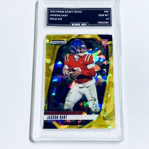 Op de afbeelding: Een JAXSON DART voetbalkaart uit de 2025 Prizm Draft Picks serie. De kaart toont een speler in een rood en wit uniform, tegen een gouden en prismatische achtergrond. De kaart is beoordeeld met GEM MT 10 en zit in een beschermende houder.
