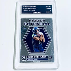 Puede incluir: Una tarjeta comercial calificada con Jaxon Smith-Njigba de los Seattle Seahawks. La tarjeta es de la serie 2023 Donruss Optic y está etiquetada como "Hidden Potential". La tarjeta está dentro de un soporte protector transparente.