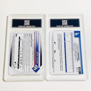 May include: Two baseball trading cards in protective cases. One card features Roki Sasaki of the Los Angeles Dodgers. The other card features Yoshinobu Yamamoto, also of the Los Angeles Dodgers. Both cards are graded by Black Box Grading Club.