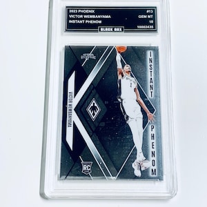 K&ouml;nnte beinhalten: Eine Basketball-Sammelkarte mit Victor Wembanyama. Die Karte hat ein schwarz-silbernes Design mit dem Text "INSTANT PHENOM" und "ROOKIE CARD". Die Karte ist in einem transparenten Schutzhalter eingeschlossen.