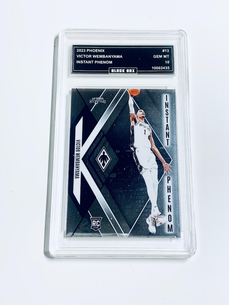 K&ouml;nnte beinhalten: Eine Basketball-Sammelkarte mit Victor Wembanyama. Die Karte hat ein schwarz-silbernes Design mit dem Text "INSTANT PHENOM" und "ROOKIE CARD". Die Karte ist in einem transparenten Schutzhalter eingeschlossen.