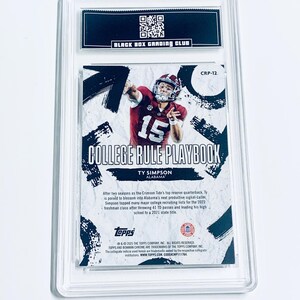 Op de afbeelding: Een voetbal trading card in een doorzichtige plastic hoes. De kaart toont een speler in een karmozijnrood shirt met nummer 15. Tekst bevat "COLLEGE RULE PLAYBOOK" en "TY SIMPSON ALABAMA". De bovenkant heeft een zwart label met "BLACK BOX GRADING CLUB".