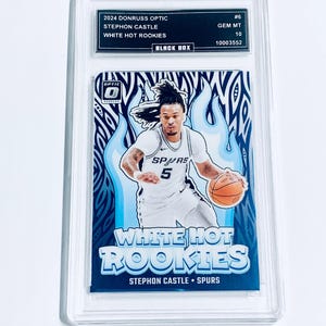 Może przedstawiać: Oceniona karta do koszyk&oacute;wki Donruss Optic 2024 z Stephonem Castle z Spurs. Karta oznaczona jest jako "White Hot Rookies" z niebiesko-białym wzorem płomienia. Karta jest zamknięta w przezroczystym uchwycie ochronnym.