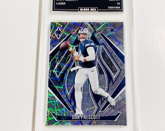 May include: A football trading card featuring Dak Prescott in a Dallas Cowboys uniform, holding a football. The card has a mosaic design with a black and silver background. The card is encased in a clear protective holder. The card reads "2024 PHOENIX DAK PRESCOTT LAZER".