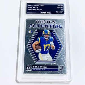 Puede incluir: Una tarjeta de fútbol Puka Nacua Hidden Potential 2023 Donruss Optic, calificada GEM MT 10, en un soporte protector. La tarjeta presenta a un jugador con uniforme azul y amarillo, con el texto "HIDDEN POTENTIAL".