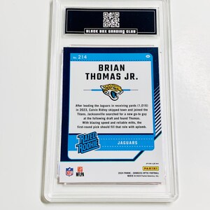 Puede incluir: Una tarjeta de f&uacute;tbol calificada con Brian Thomas Jr. La tarjeta tiene un dise&ntilde;o azul y verde azulado con el texto "RATED ROOKIE" y "JAGUARS". La tarjeta est&aacute; encerrada en un soporte protector transparente con el texto "BLACK BOX GRADING CLUB".