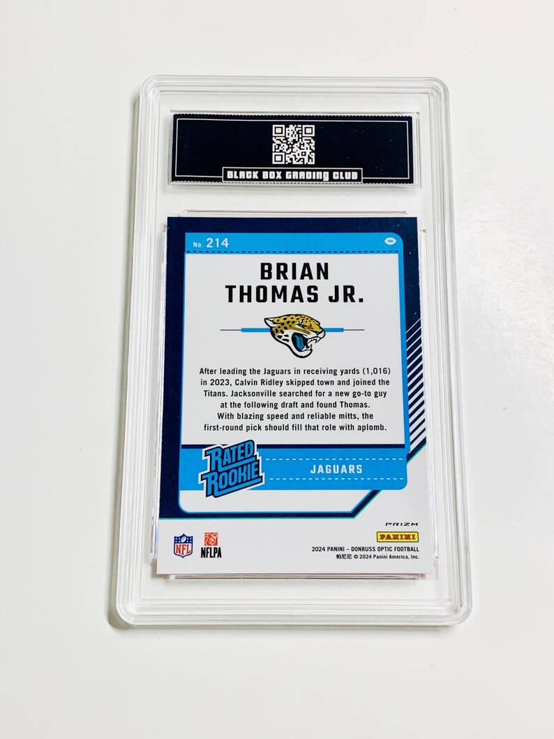 Puede incluir: Una tarjeta de f&uacute;tbol calificada con Brian Thomas Jr. La tarjeta tiene un dise&ntilde;o azul y verde azulado con el texto "RATED ROOKIE" y "JAGUARS". La tarjeta est&aacute; encerrada en un soporte protector transparente con el texto "BLACK BOX GRADING CLUB".