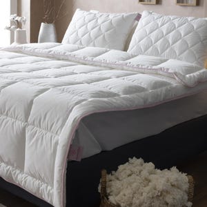 May include: A white quilted comforter and pillows with pink trim are displayed on a bed. A woven basket filled with fluffy white material sits at the foot of the bed. The bedding is in a bedroom setting.