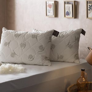 May include: Two white, quilted pillows with a floral pattern rest on a bed. A decorative lamp and framed artwork are in the background. A glass bottle and a small pile of white material are on the bed.