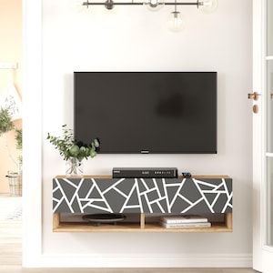 May include: A floating wall mounted TV stand with a wood grain finish and a black and white geometric pattern on the front. The stand has two shelves, one for a TV and one for storage. The stand is mounted on a white wall.