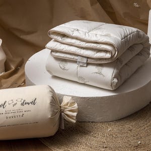 May include: A cream-colored wool duvet and a rolled-up duvet in a matching fabric. The rolled duvet is labeled "wool duvet" with text and illustrations. The folded duvet is on a white pedestal, with a neutral background.