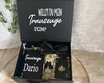 May include: A black gift box with the German text "WILLST DU MEIN Trauzeuge SEIN?" on the lid. Inside, a flask, bow tie, shot glass with a mustache, and playing cards. The flask is personalized with the name "Dario".