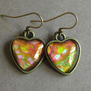 May include: A pair of heart-shaped earrings with a bronze-colored frame. The colorful design inside the heart features swirls of orange, yellow, pink, and green. The earrings have a hook closure.