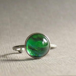 May include: A silver ring featuring a round, vibrant green gemstone. The gemstone has a swirling pattern of dark green and black. The ring's band is thin and made of silver-colored metal. The setting around the stone is also silver.