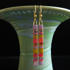 May include: Gold-tone dangle earrings with a vertical design. The earrings feature a series of colorful rectangular beads in shades of lavender, yellow, orange, and red. The earrings are displayed against a green vase.