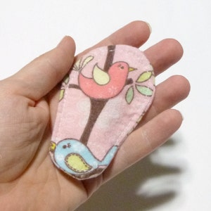 May include: A pink fabric item with a bird print, held in a hand. The print features two birds, one pink and one blue, perched on a brown tree branch with green leaves. The item has a stitched edge.