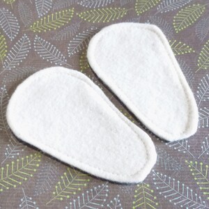 May include: Two white, oval-shaped felt insoles with a stitched border. The insoles are positioned on a brown fabric with a repeating pattern of green and white leaf-like designs.