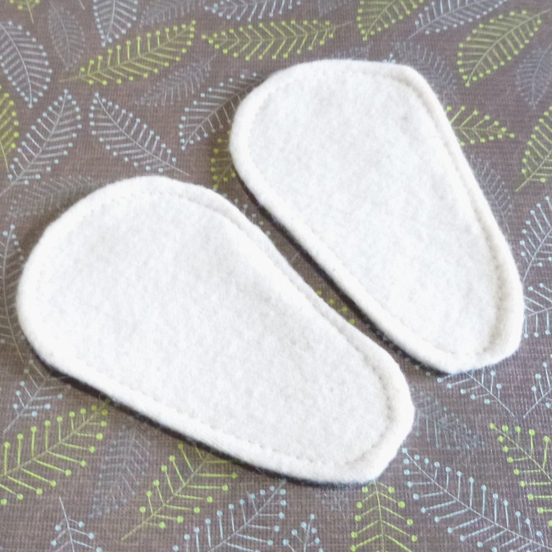May include: Two white, oval-shaped felt insoles with a stitched border. The insoles are positioned on a brown fabric with a repeating pattern of green and white leaf-like designs.